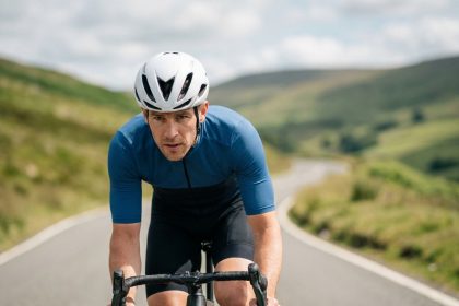 Cycliste portant un casque vélo de route aérodynamique blanc sur une route asphaltée avec des collines verdoyantes en arrière-plan